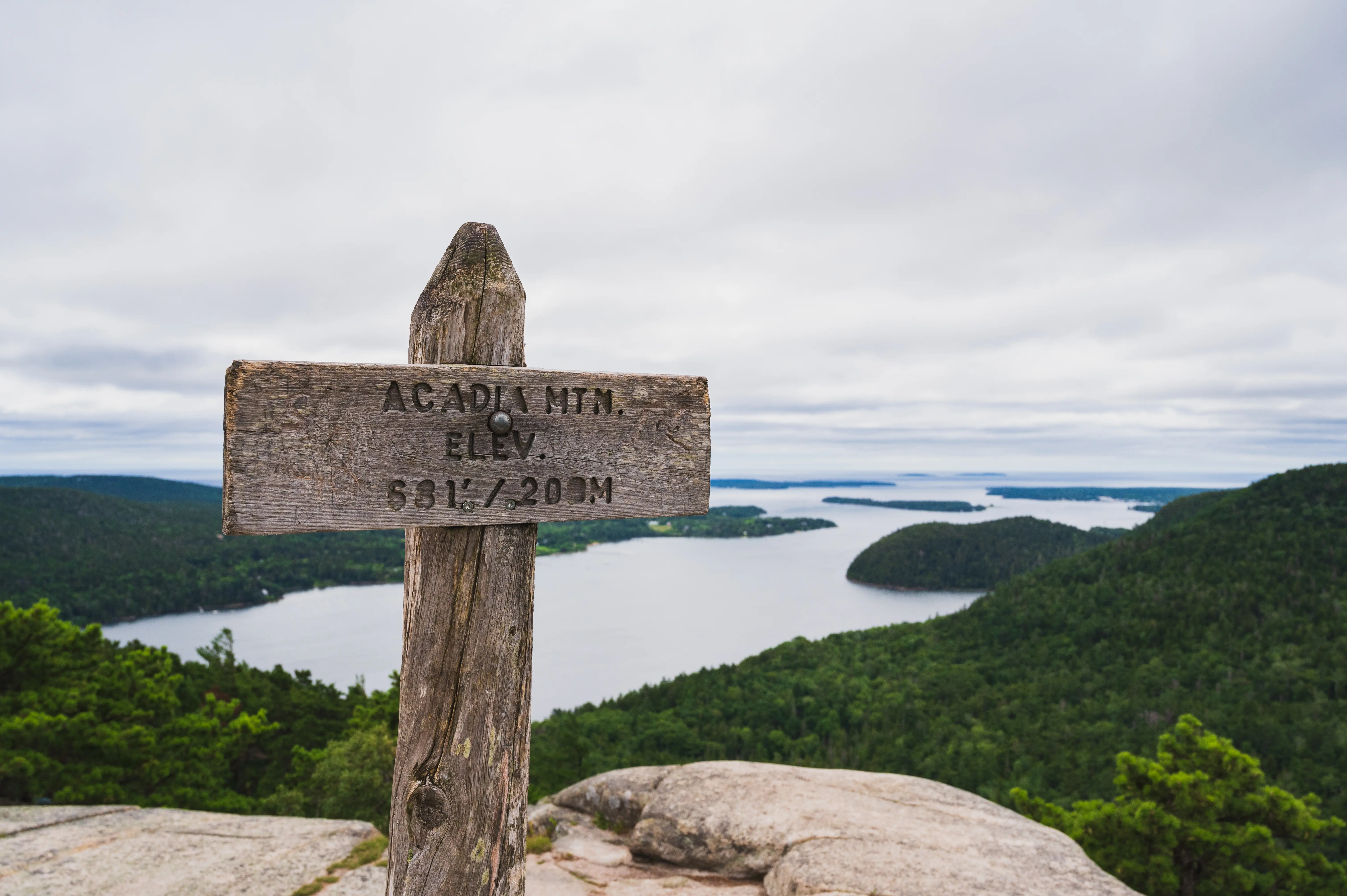 a wooden sign reads 'Acadia Mountain, Elevation 681 feet' while islands and trees flow out into the ocean beyond